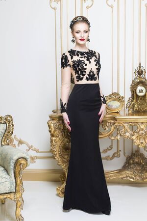 Beautiful blonde royal woman standing near retro table in gorgeous luxury dress looking in camera. Indoorの写真素材