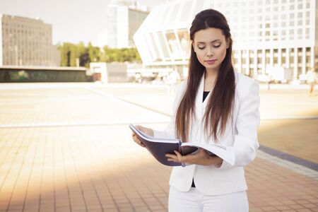 Beautiful brunette business woman in white suit with folder of documents in her hands reading outdoors. copy spaceの写真素材