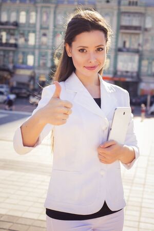 Beautiful brunette business woman in white suit with tablet in her hands outdoors showing thumb upの写真素材