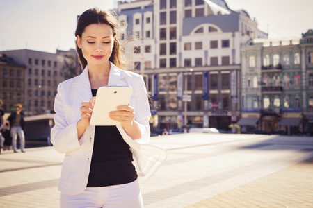 Beautiful brunette business woman in white suit working on a tablet in her hands outdoors. copy spaceの写真素材