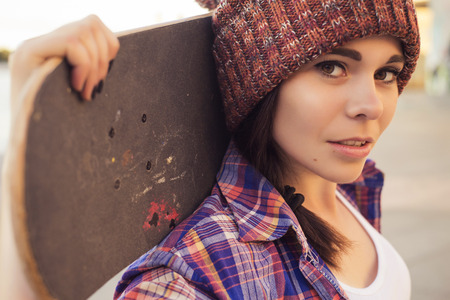 Brunette teenage girl in hipster outfit (jeans shorts, keds, plaid shirt, hat) with a skateboard at the park outdoorsの写真素材