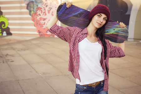 Brunette teenage girl in hipster outfit (jeans shorts, keds, plaid shirt, hat) with a skateboard at the park outdoors. Copy spaceの写真素材
