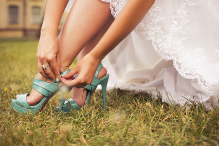 Beautiful bride preparing to get married in white dress and fasten her  blue shoes on the grass in the park alone. outdoors. copy spaceの写真素材