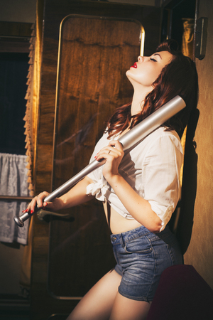 Beautiful sexy brunette retro woman with pin-up hairstyle and make-up in a shorts and shirt with a bat in her hands in vintage interior. Indoors. Retro tonedの写真素材