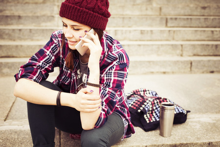 Hipster woman in plaid shirt and hat talking on the phone sitting on the steps in european city. Generation Z conceptの写真素材