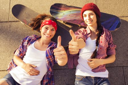 Two Brunette teenage girls friends in hipster outfit (jeans shorts, keds, plaid shirt, hat) with a skateboard at the park outdoors.の写真素材