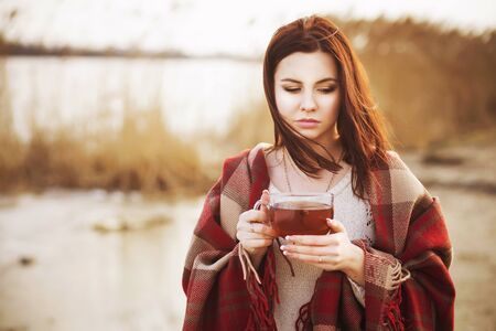 woman in plaid blanket on a lake drinks teaの写真素材