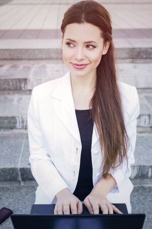 Beautiful brunette business woman in white suit with notebook on her lap, typing, working outdoors. Copy spaceの写真素材