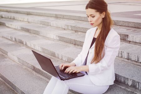 Beautiful brunette business woman in white suit with notebook on her lap, typing, working outdoors. Copy spaceの写真素材