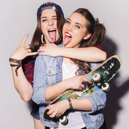 Two beautiful brunette women (girls) teenagers spend time together having fun, make funny faces. Casual hipster outfit, jeans jacket and plaid shirt, with a skateboardの写真素材