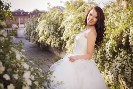 Beautiful brunette woman bride in a garden park in white wedding dress, curly hairstyle and a smile. warm weather, outdoors. copy spaceの写真素材