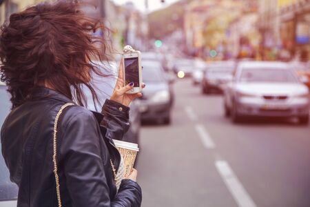 Brunette beautiful stylish caucasian woman in casual outfit on a walk on european city street photographing on her phone. copy spaceの写真素材