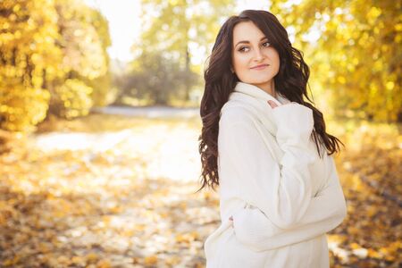 Beautiful caucasian brunette young woman in warm autumn day at park. Yellow leaves and sunshine. Fall season. Woman in white sweater, smiling. Copy spaceの写真素材