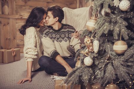 Beautiful brunette caucasian romantic loving couple in cozy warm sweaters in the cabin on a christmas new year eve, smiling, kissing, having fun with each other. Dim dark night light, toned imageの写真素材