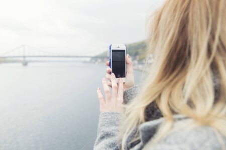 Blonde beautiful stylish caucasian woman in casual outfit on a walk in european city street  photographing view: bridge and river on her phone. copy space. focus on the phoneの写真素材