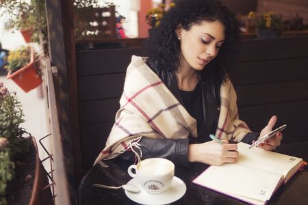 Beautifil brunette caucasian woman in leather jacket and plaid sitting on a cafe drinking coffe and working on her phone and notebook. cold weatherの写真素材