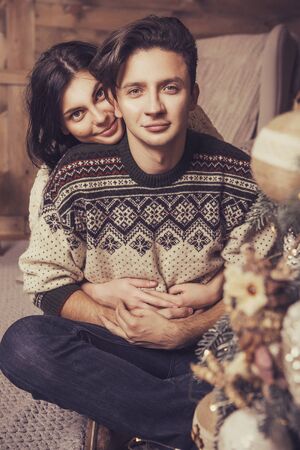 Beautiful brunette caucasian romantic loving couple in cozy warm sweaters in the cabin on a christmas new year eve, smiling, kissing, having fun with each other. Dim dark night light, toned imageの写真素材