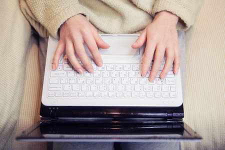 Woman sitting on a couch with a laptop on her knees, working. Writing on her blog, typing messages in social media. Soft light, indoorsのeditorial素材