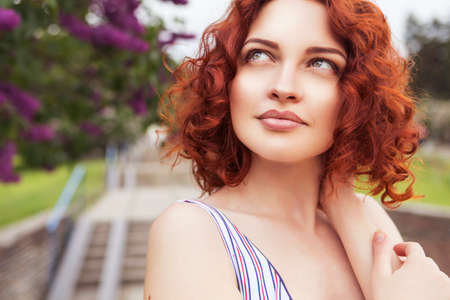 Beautiful red haired woman with fresh flawless skin and curly hairstyle in dress, smiling cheerfully...