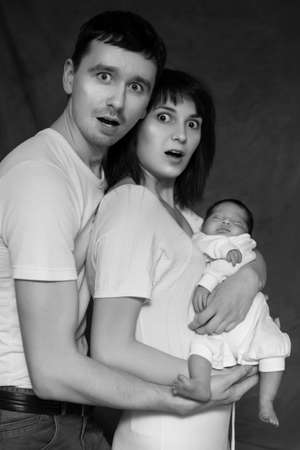 Little infant baby boy sleeping laying on mothers and fathers arms. Mother and father shocker scared. Neutral black background, black and white picture. All in white clothes. Happy familyの写真素材