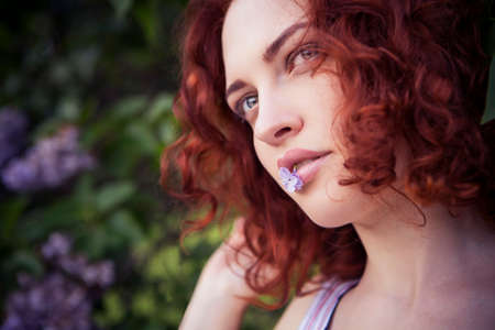 Beautiful red haired woman with fresh flawless skin and curly hairstyle in dress in park near flowers.の写真素材