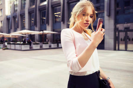 Beautiful rich casual blonde stylish fashion business woman with a phone in her hand working in pink jacket and black trousers. European buildings and street on a background. Copy spaceの写真素材