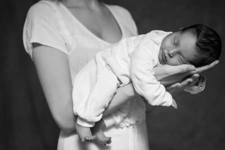 Little infant baby boy sleeping laying on mothers arms. Focus on a boy. Neutral black background, black and white picture. All in white clothes. Happy familyの写真素材