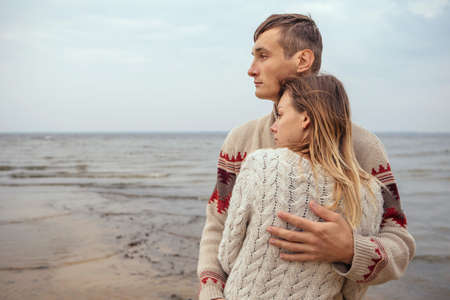 Happy thoughtful couple standing on a rock beach near sea hugging each other in cold foggy cloudy autumn weather. Copy spaceの写真素材