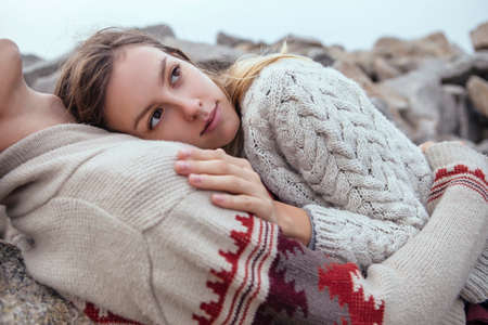 Happy thoughtful couple sitting on a rock beach near sea hugging each other in cold foggy cloudy autumn weather. Copy spaceの写真素材