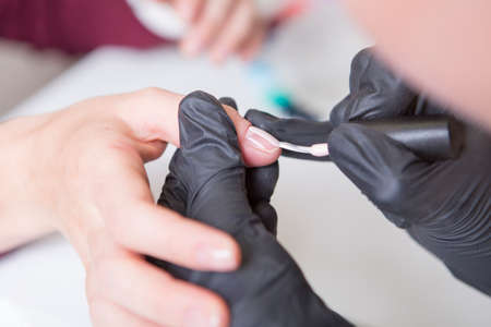 Nail master in rubber gloves apply gel polish shellac on a woman hand finger nail. Healthcare beauty cosmetic spa procedure in a salonの写真素材