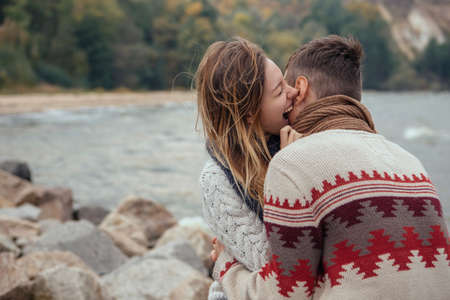 Happy thoughtful couple sitting on a rock beach near sea hugging each other in cold foggy cloudy autumn weather. Copy spaceの写真素材