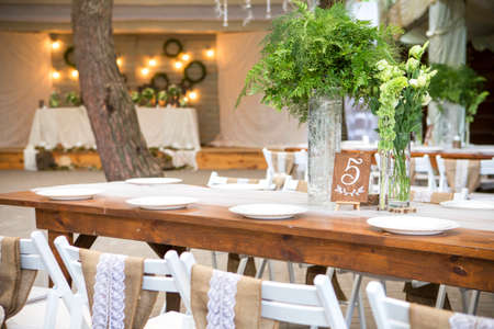 Fresh eustoma roses bouquet composition on a table in a glass vase. Wedding bridal decor. Dim dark sunset light. Romantic mood. Chairs with rustic styled decotarion in a shape of heartの写真素材