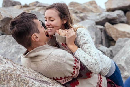 Happy thoughtful couple sitting on a rock beach near sea hugging each other in cold foggy cloudy autumn weather. Copy spaceの写真素材