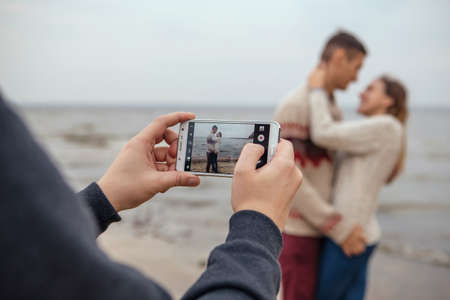 Happy thoughtful couple standing on a rock beach near sea hugging each other in cold foggy cloudy autumn weather. Man photograph them on a phone. Copy spaceの写真素材