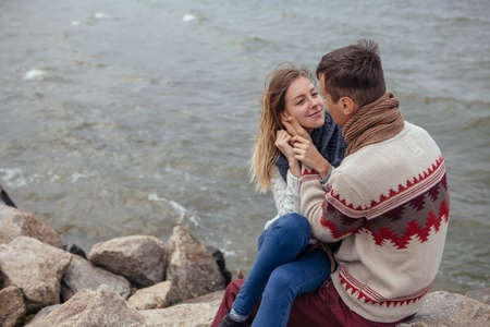 Happy thoughtful couple sitting on a rock beach near sea hugging each other in cold foggy cloudy autumn weather. Copy spaceの写真素材