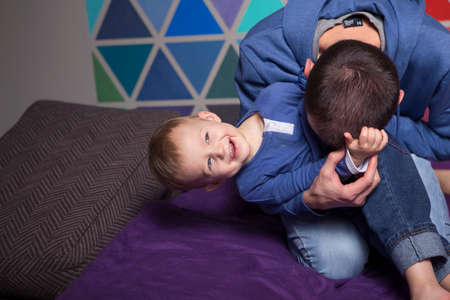 Cute caucasian ordinary family in casual clothes at home sitting on a couch. Lifestyle portrait of dad and two years old child son playing together, have fun. Colorful backgroundの写真素材
