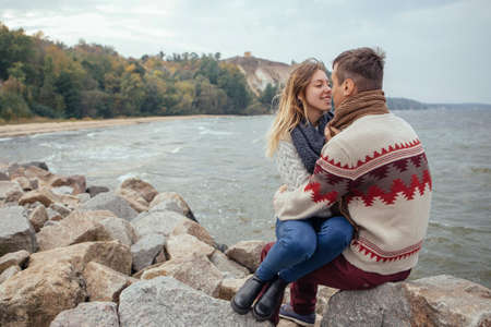 Happy thoughtful couple sitting on a rock beach near sea hugging each other in cold foggy cloudy autumn weather. Copy spaceの写真素材
