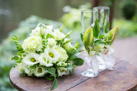 Bouquet of eustoma roses and glassess decorated with flowers lay on wooden table on a wedding ceremony. Soft day light, outdoor in gardenの写真素材