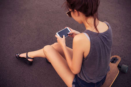 Young beautiful brunette caucasian sexy adult woman outdoors in city, having fun skateboarding (on longboards). Typing message on her phone. Summer weatherの写真素材