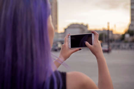 Beautiful brunette woman with colored (purple violet blue) hair in dark black slim dress outdoor in the european city, smiling, typing, playing, make photo on her phoneの写真素材