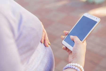 Beautiful brunette caucasian pregnant woman on summer walk outdoors, holding her belly in a soft, sensual, maternity way. In casual jeans dress and bride hairstyle typing message on phone. Copy spaceの写真素材