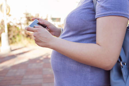 Beautiful brunette caucasian pregnant woman on summer walk outdoors, holding her belly in a soft, sensual, maternity way. In casual jeans dress and bride hairstyle typing message on phone. Copy spaceの写真素材