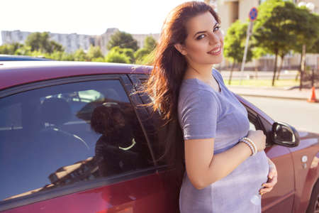 Beautiful brunette caucasian pregnant woman on summer walk outdoors, holding her belly in a soft, sensual, maternity way. In casual jeans dress and bride hairstyle, standing near red car. Copy spaceの写真素材