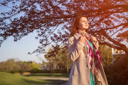 Cute ordinary caucasian brunette woman in casual outfit walking at the nature park. Cold spring (autumn) weather, warm coat on a girl. Sunset. Copy spaceの写真素材