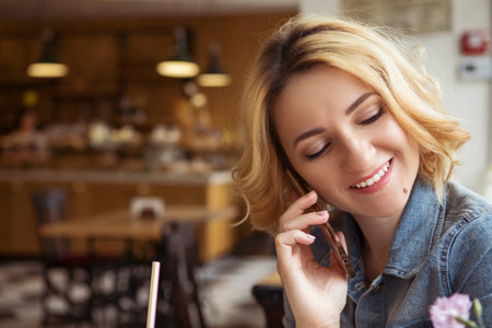 Blonde (with dark roots) caucasian woman in casual summer outfit at the cafe. Gray dress and jeans jacket. Woman got natural day makeup and curly hairstyle. She talks on her phone, working, make callsの写真素材