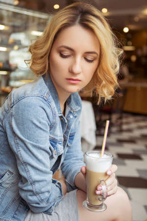 Blonde (with dark roots) caucasian woman in casual summer outfit at the cafe. Gray dress and jeans jacket. Woman got natural day makeup and curly hairstyle. She talks on her phone, working, make callsの写真素材