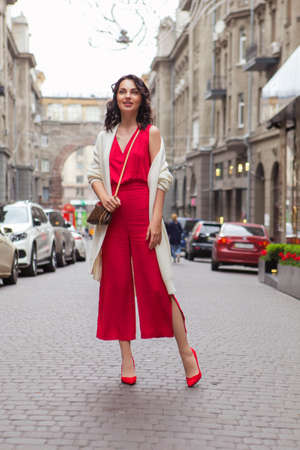 Beautiful brunette caucasian business woman sitting in cafe on a coffee break in red casual but a bit classic dress and white cardigan, smile, talking on the phone. Copy space. Evening, sunset lightの写真素材