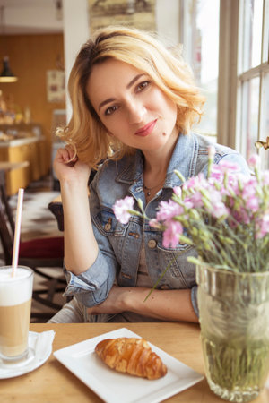 Blonde (with dark roots) caucasian woman in casual summer outfit at the cafe. Gray dress and jeans jacket. Woman got natural day makeup and curly hairstyle. She talks on her phone, working, make callsの写真素材