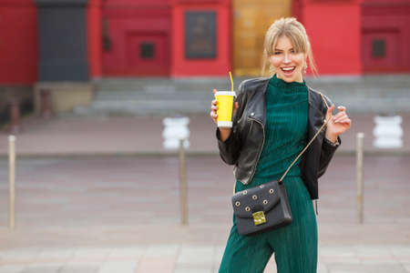 Beautiful rich luxury looking blonde caucasian business woman in casual green outfit standing on a european streets, holding coffee and purse. Daylight, outdoors.の写真素材