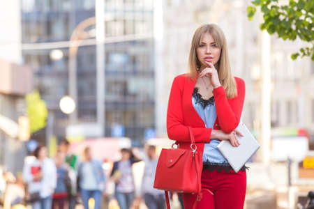 Beautiful rich luxury looking blonde caucasian business woman in smart-casual red outfit standing on a european streets near cars, holding tablet, distant working. Daylight, outdoors.の写真素材